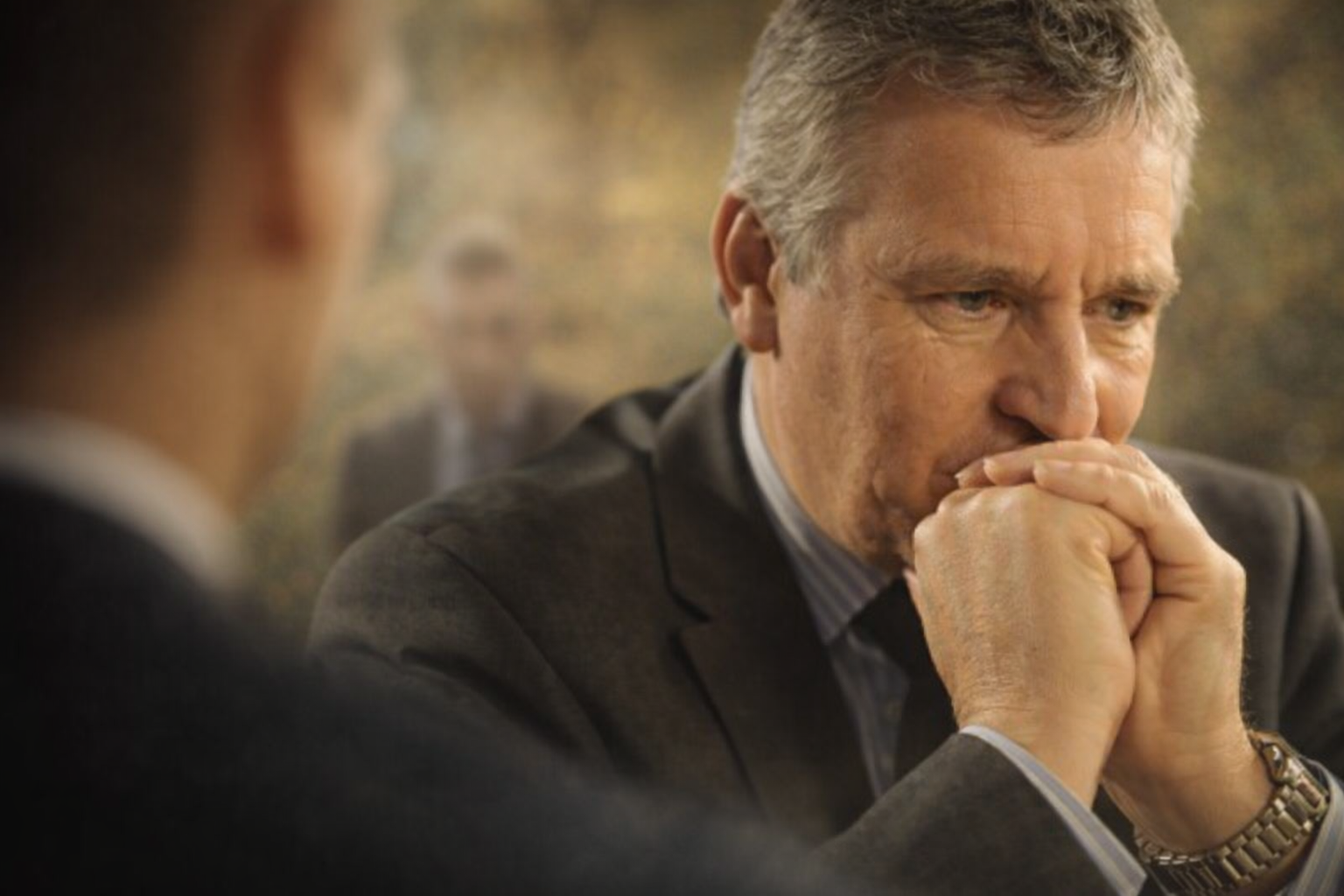 Homme en costume, assis en réunion, les mains jointes devant la bouche dans une posture de réflexion et de tension. Son regard est concentré et préoccupé, tourné vers un interlocuteur partiellement visible au premier plan à gauche, suggérant un échange im